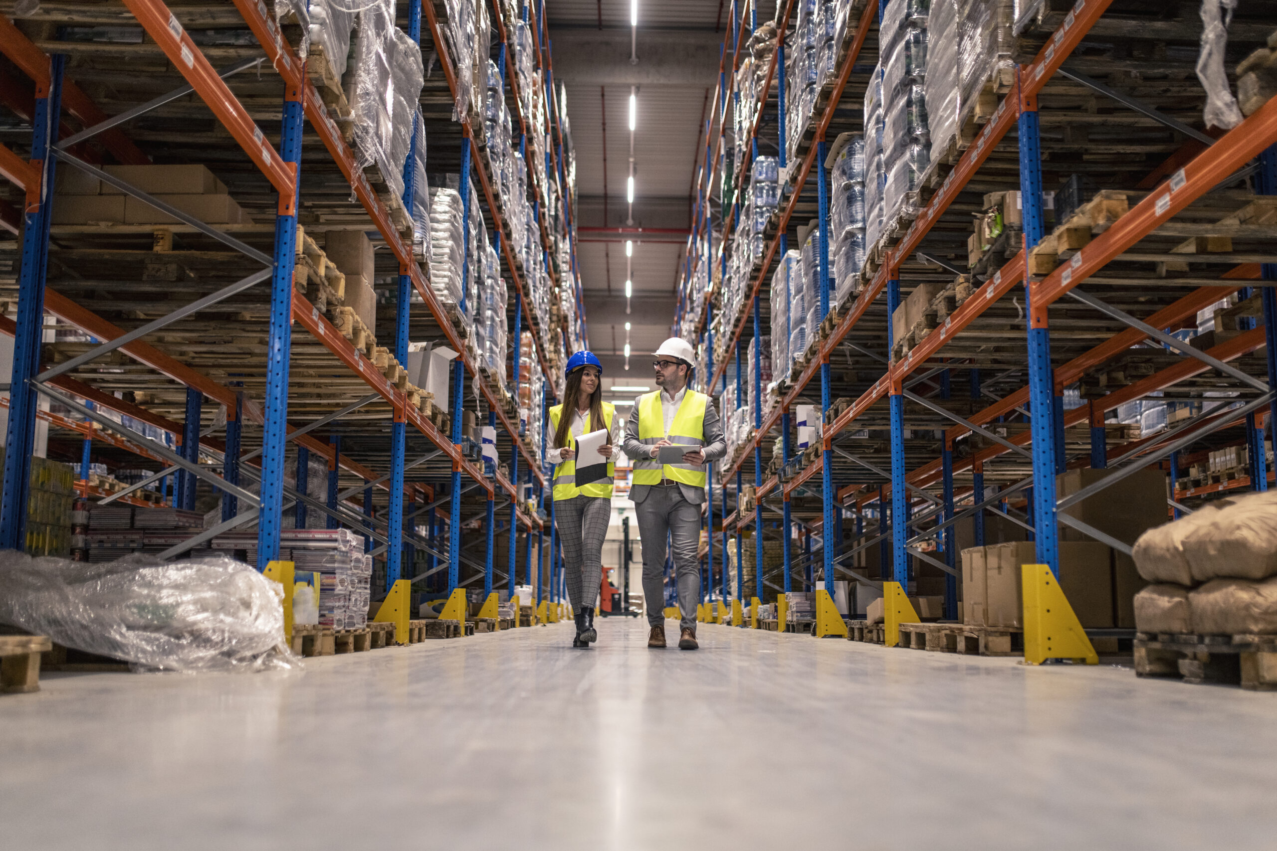 workers with hardhats reflective jackets walking through big warehouse aisle checking goods condition scaled